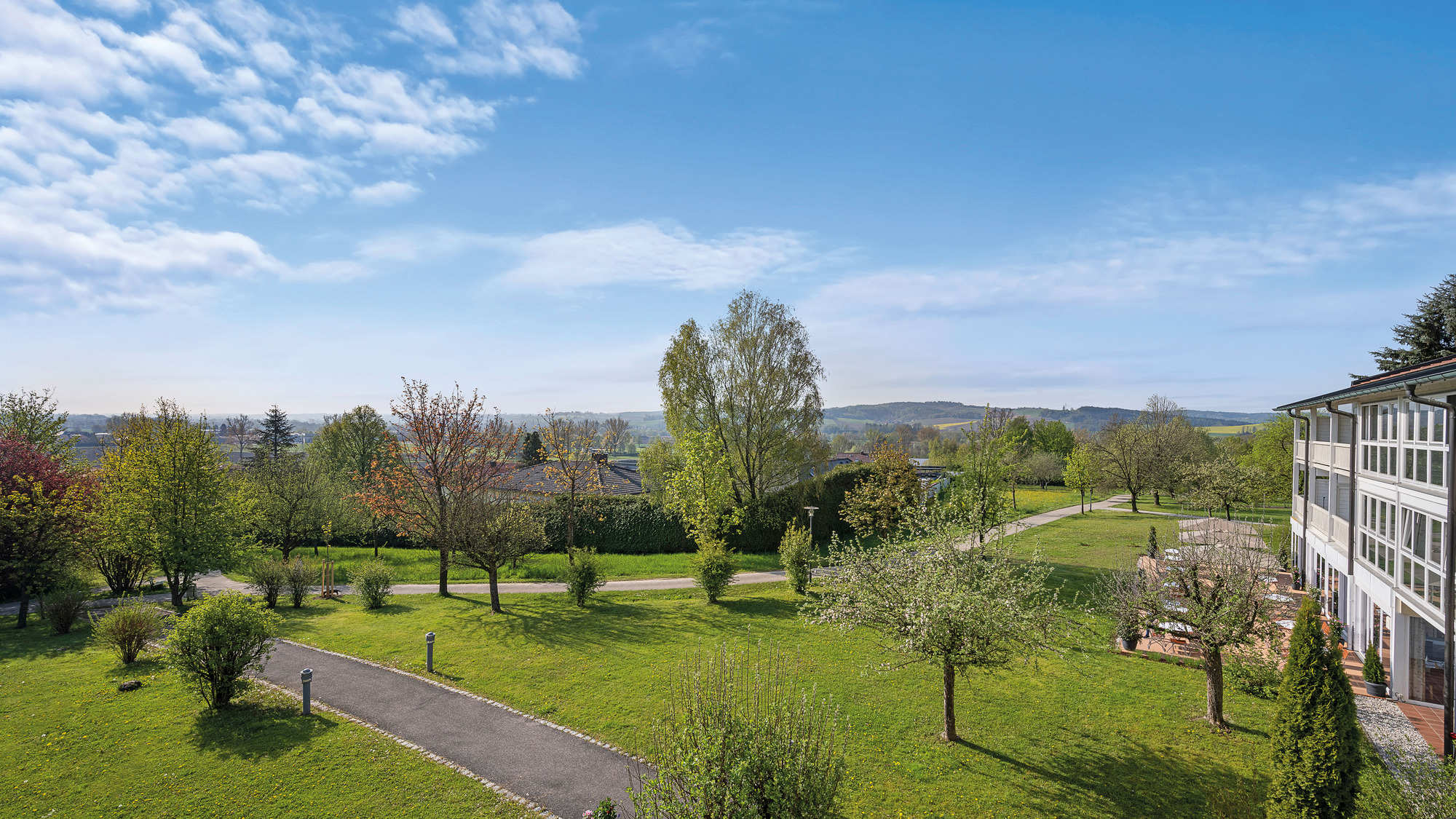 Gepflegter Garten mit Bäumen, Wegen und Blick auf Häuser bei klarem Himmel.