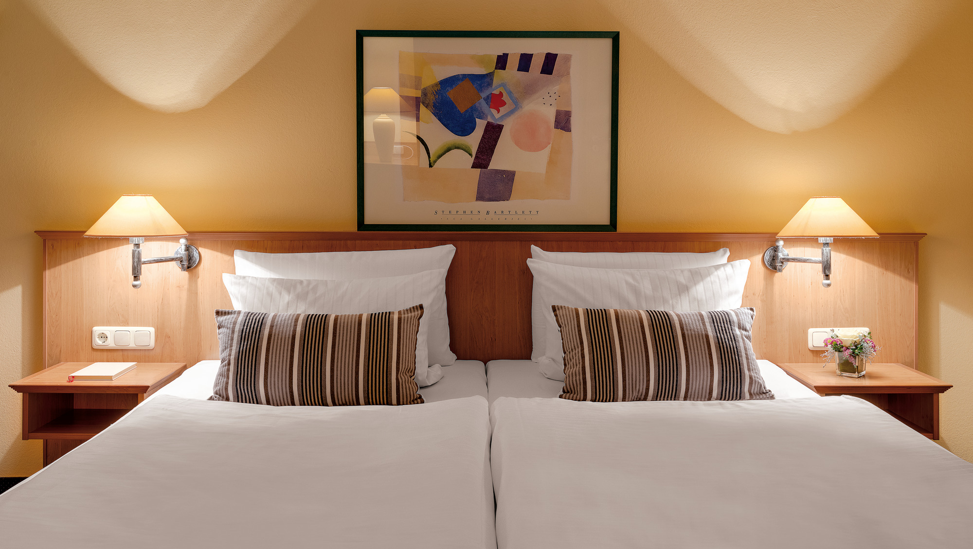 Zwei Einzelbetten mit weißer Bettwäsche, Holzmöbeln, Wandlampen und Blumen.