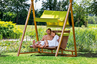 Zwei Personen entspannen auf Schaukel im Garten, umgeben von grünen Bäumen.