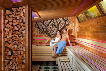 Zwei Personen in Sauna mit Holzdekor und Baum-Motiv, warmes Licht, entspannte Stimmung.