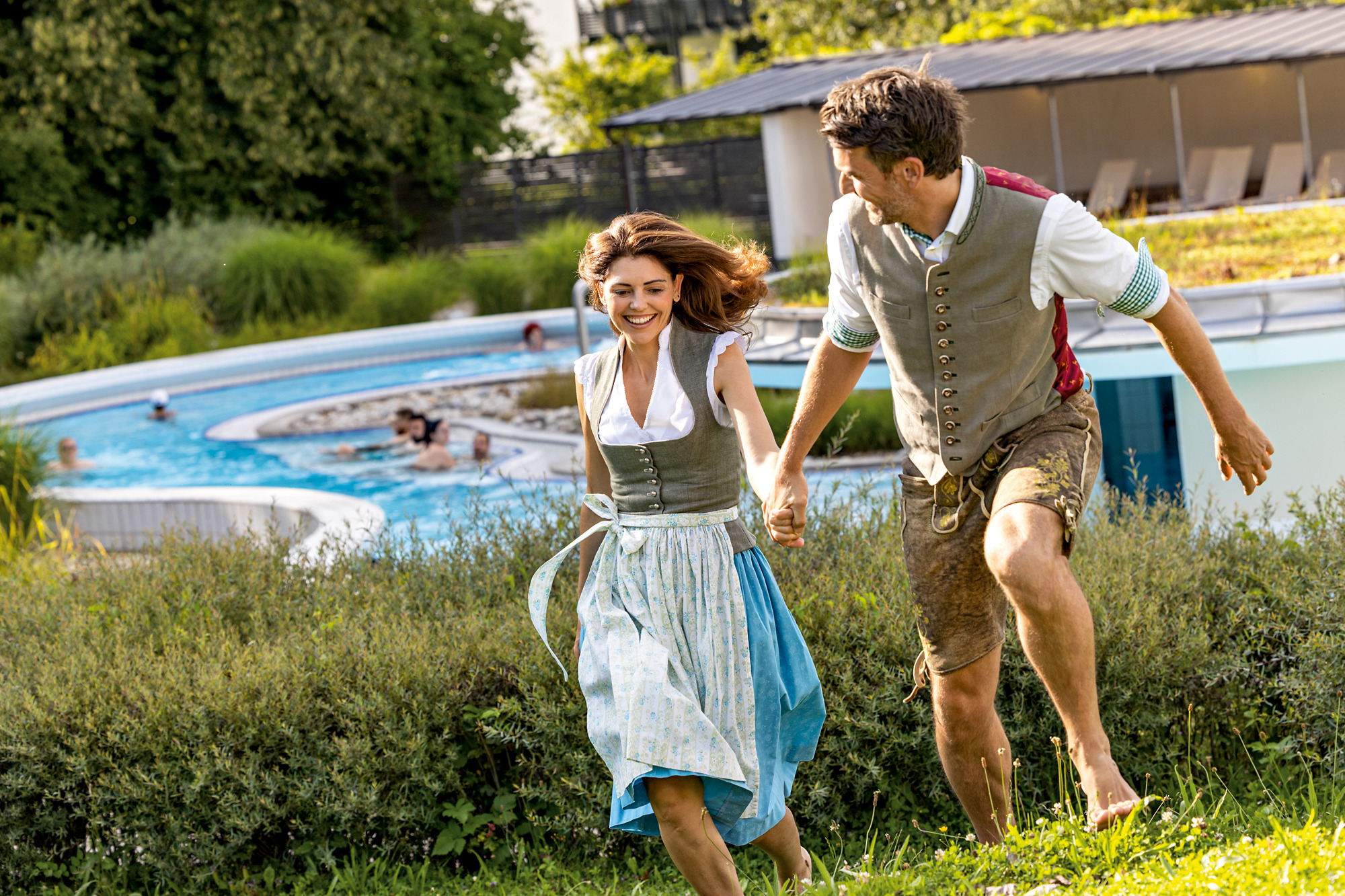 Zwei Personen in Tracht laufen Hand in Hand über Wiese, Pool im Hintergrund.